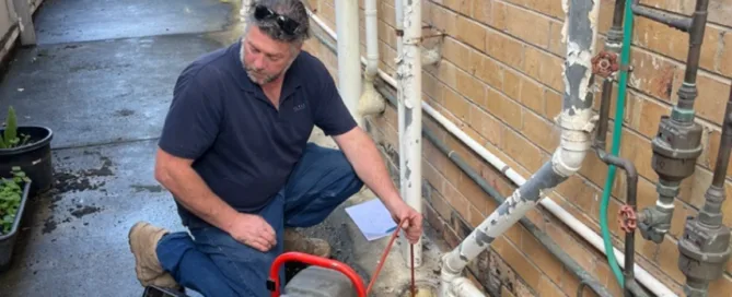 A man in a blue collared shirt and jeans measures an outdoor pipe using a measuring tool. He is standing in an alley next to a plumbing machine with red handles and a red pipe connected to the main pipe.
