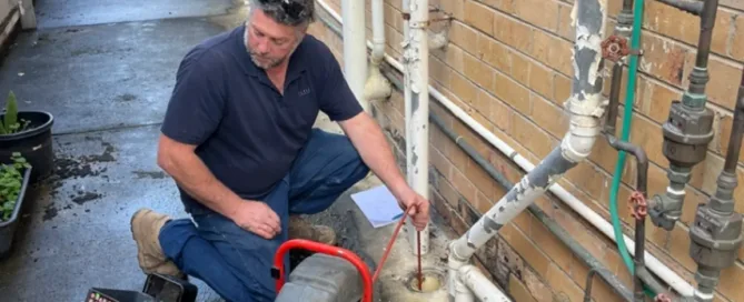 A man in a blue collared shirt and jeans measures an outdoor pipe using a measuring tool. He is standing in an alley next to a plumbing machine with red handles and a red pipe connected to the main pipe.