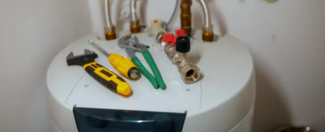 Close-up of a residential water heater with valves and assorted plumbing tools on top, ready for maintenance in a utility room.
