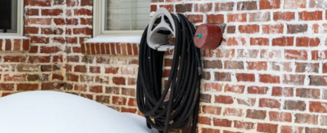 Winterize outdoor plumbing: insulated outdoor faucet and garden hose on a brick wall surrounded by snow to prevent freezing and pipe damage.