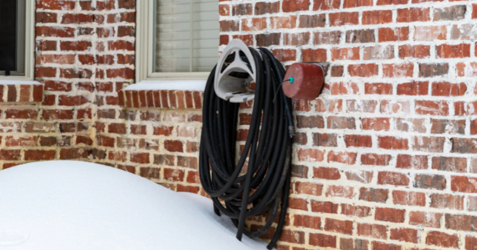 Winterize outdoor plumbing: insulated outdoor faucet and garden hose on a brick wall surrounded by snow to prevent freezing and pipe damage.