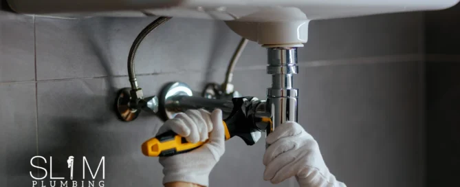 Prevent Clogged Drains Before They Start - Slim Plumbing plumber tightening under-sink drain pipe with wrench to keep drains clear